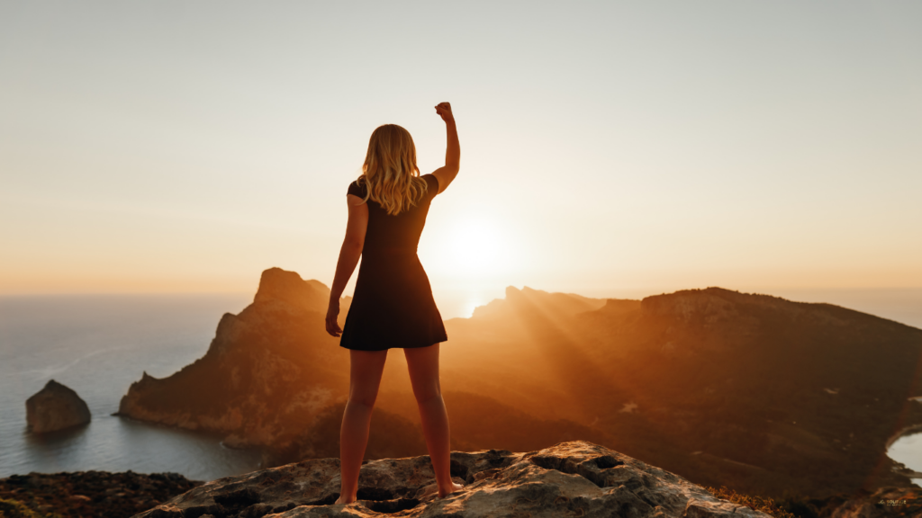 Victory over chronic pain: Interpretation Matters. A young women standing on a rock looking over the ocean with arm up in the air in victory.